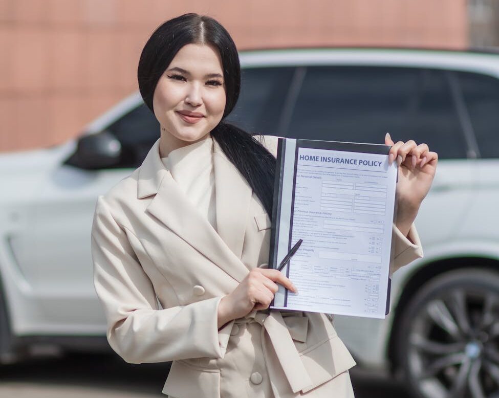 Femme souriante en blazer beige détenant un document de police d'assurance habitation en extérieur.