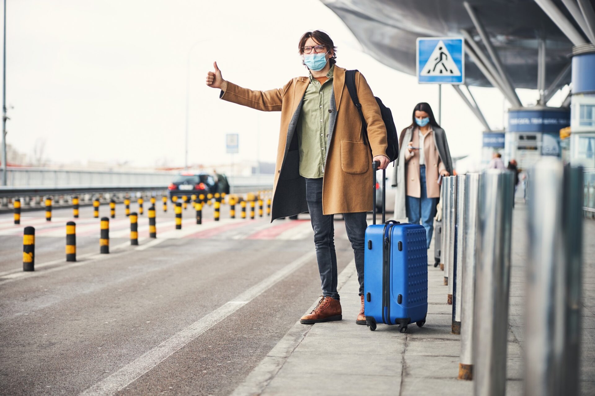 Good Gentleman with Traveler Suitcase portant un masque de visage protecteur tout en faisant un geste de pouce et en essayant d'arrêter le taxi dans la rue